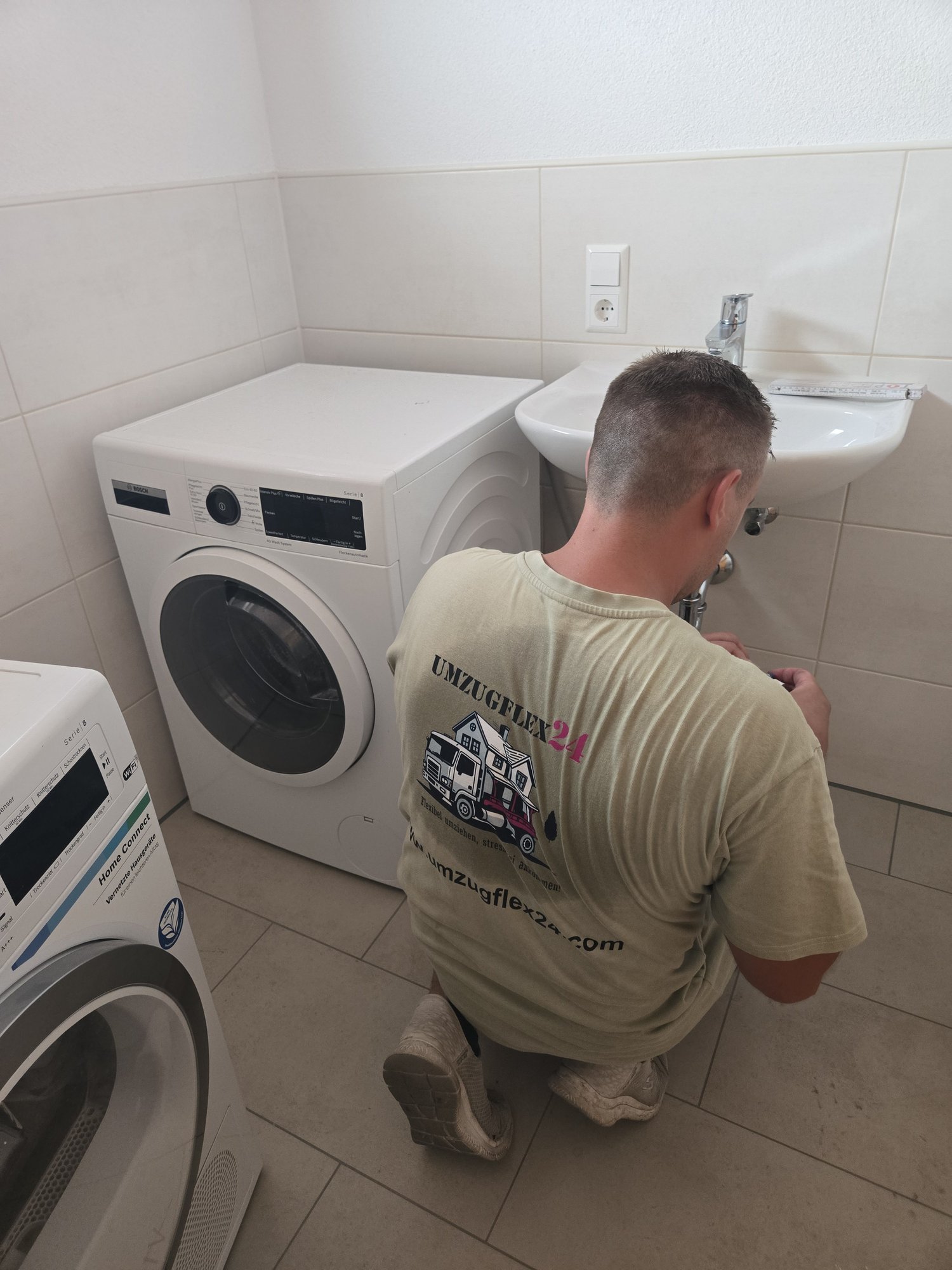 Monteur von Umzugflex24 schließt in der neuen Wohnung die Waschmaschine an, daneben steht der Trockner